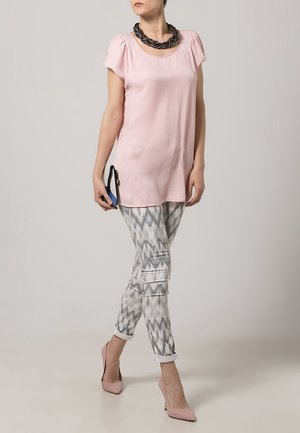 Mujer con camiseta de manga corta rosa claro, pantalones grises y blancos con estampado, tacones rosas y collar trenzado grueso negro, sosteniendo un par de zapatos.