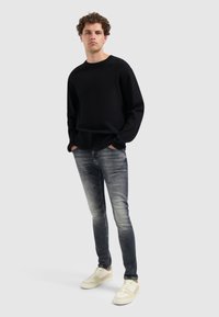 Schwarzer Strickpullover, ausgebleichte graue Skinny Jeans und beige Sneakers. Das Model steht mit den Händen in den Taschen vor einem neutralen Hintergrund.