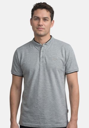 Homme portant un t-shirt henley à manches courtes gris clair avec trois boutons et des manches bordées de noir, debout devant un fond uni.