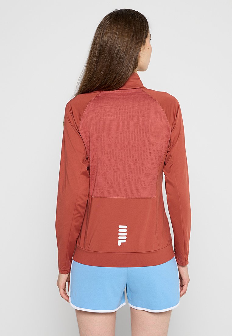 Chaqueta deportiva de manga larga en óxido con parte trasera texturizada, con un logo blanco cerca del dobladillo. Combinada con pantalones cortos azul claro.