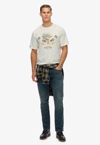 Lys grå grafisk t-shirt med drageprint og "Superdry" logo, parret med mørke jeans og en ternet skjorte bundet om livet. Brune sko.