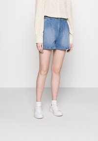 Farkkushortsit vaaleansinisinä, joissa on elastinen vyötärö, rento istuvuus ja riiputettu helma. Yhdistetty pitkähihhaisen, pilkullisen paidan ja valkoisten lenkkareiden kanssa.