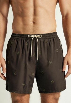 Torace e parte superiore delle gambe di un uomo che indossa pantaloncini da bagno scuri con piccole stampe di paperelle gialle di gomma e coulisse bianca.