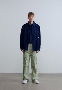 Πουκάμισο ναυτικού με κουμπιά πάνω από σκούρο τοπ, συνδυασμένο με ανοιχτό πράσινο cargo παντελόνι με πλαϊνές τσέπες. Το μοντέλο φορά λευκά αθλητικά παπούτσια.