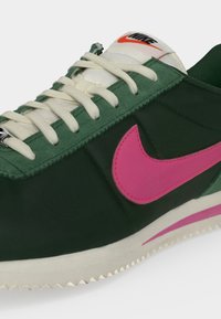Green Nike sneaker with white laces, a pink swoosh logo on the side, and an orange Nike logo on the white tongue.