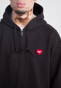 Black zip-up hoodie made of cotton with textured fabric, featuring a red heart patch with "Carhartt WIP" logo and drawstring detail.