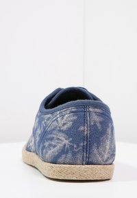 Chaussure en tissu bleu à enfiler avec des motifs de feuilles blanches décolorées et une semelle beige tissée, vue de dos sur une surface blanche.