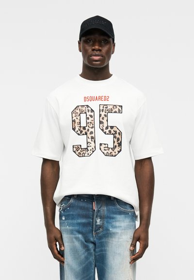 Άνδρας που φοράει λευκό T-shirt DSQUARED2 με αριθμό 95 σε λεοπάρ σχέδιο, μπλε τζιν με φθορές και μαύρο καπέλο με την επιγραφή «VOLTAIRE».