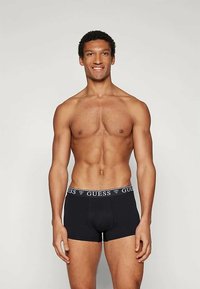 Črne bokserice z teksturiranim pasom, ki vsebuje logo "GUESS". Prilagojen kroj s gladkim materialom in kratkim dizajnom nogavic.
