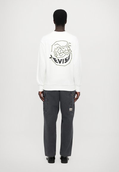 Hvid sweatshirt med "EVISU" logo og grafisk design på ryggen, parret med mørkegrå cargo bukser med patchdetaljer og lommer.