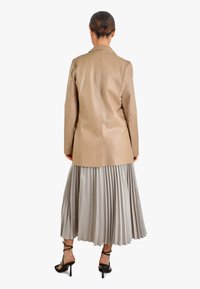 Beżowa skórzana marynarka o dopasowanym kroju, zestawiona z jasnoszarą plisowaną spódnicą midi i czarnymi sandałami na paski, widziana od tyłu.