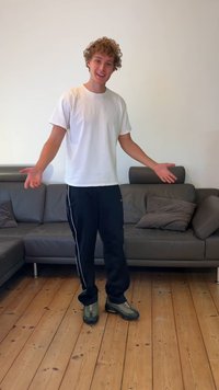 Sportieve schoen in grijs en zwart, met een getextureerd oppervlak en afgeronde neus. De persoon staat op een houten vloer vlak bij een grijze bank.