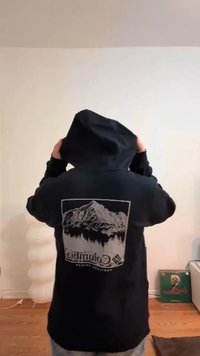 Camisola com capuz preta com um gráfico de montanha e árvores nas costas, apresentando um capuz com cordão e punhos canelados. Textura suave do tecido.