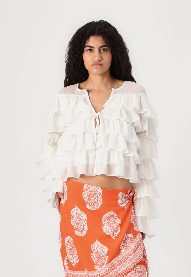 Hvit bluse med rysjer og knytning foran, lagdelt design og semi-transparent tekstur, sammen med et oransje mønstret skjørt med paisleymønster.