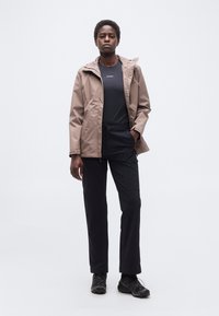 Γυναίκα όρθια που φοράει ανοιχτό καφέ μπουφάν, μαύρο μπλουζάκι, μαύρο παντελόνι και μαύρα αθλητικά παπούτσια μπροστά σε απλό λευκό φόντο.