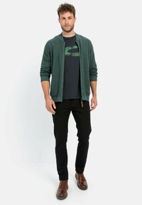 Grüne Zip-Up-Strickjacke mit strukturiertem Gestrick, kombiniert mit einem schwarzen T-Shirt mit grünem Logo. Schwarze Slim-Fit-Hosen und braune Schuhe.