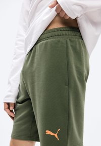 Άτομο σηκώνει λευκό πουκάμισο για να δείξει πράσινα σορτς Puma με ελαστική μέση και πορτοκαλί λογότυπο Puma στο αριστερό μηρό.