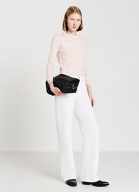 Frau steht in einem hellrosa gesprenkelten Pullover, hochgeschnittenen weißen Hosen, schwarzen spitzen Schuhen und hält eine große schwarze Clutch.