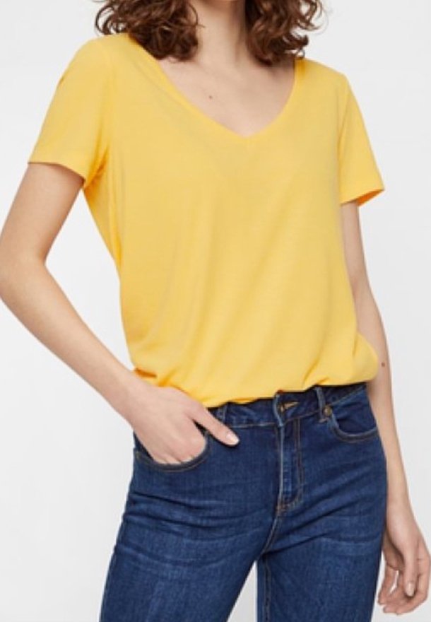 Kvinna som bär en gul V-ringad kortärmad tröja in stoppar i blå denimjeans med en hand i fickan.