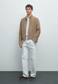 Beige Wildleder Bomberjacke mit Stehkragen, weißem Button-Down-Hemd, weißen Hosen und braunen Slippern mit strukturiertem Boden.
