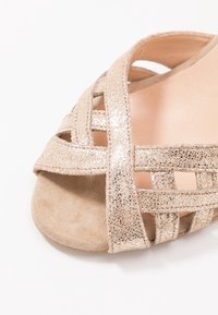 Sandal con punta aperta caratterizzata da cinturini metallici incrociati in un tono oro chiaro, posata su una morbida soletta beige. Il materiale combina texture lucide e scamosciate.