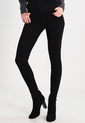 Person iført sorte skinny bukser og sorte ankelstøvler med hæl, stående med det ene ben let bøjet.