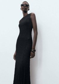 Robe maxi noire en tricot avec une silhouette ajustée. Présente un design asymétrique à une épaule et un tissu texturé. Le modèle porte des lunettes de soleil et des bracelets en perles.