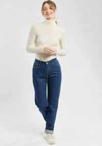 Hellbeige Rollkragenpullover kombiniert mit dunkelblauen, hoch taillierten Jeans, mit aufgerollten Manschetten und weißen Sneakers. Einfaches, tailliertes Design.