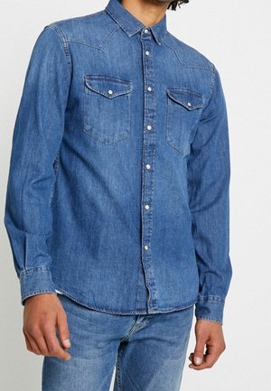 Uomo che indossa una camicia di denim blu a maniche lunghe con bottoni sul davanti e due tasche sul petto abbinata a jeans blu.