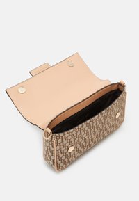 Malá obdélníková béžová crossbody taška s monogramem, otevřeným klopou a zlatými magnetickými patentkami, odhalující černou vnitřní látku.