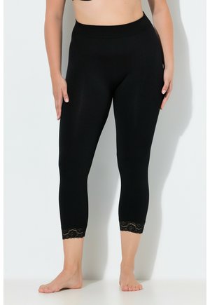 Kobieta ubrana w czarne, krótkie legginsy z wysokim stanem z koronkowym wykończeniem przy kostkach, stojąca boso na białym tle.