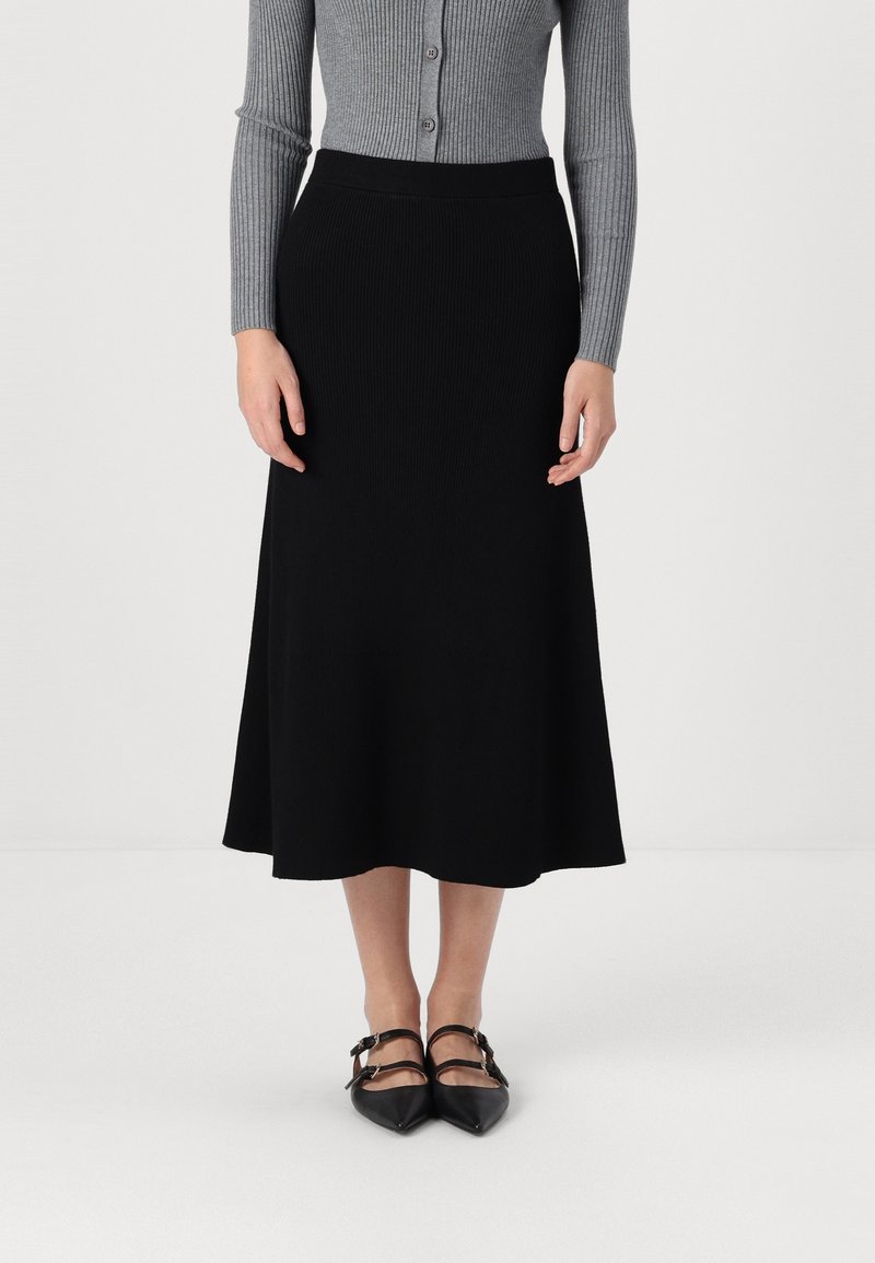 Femme portant une jupe midi noire, un haut gris côtelé à manches longues boutonné et des chaussures Mary Jane noires à bout pointu, debout sur un fond blanc.