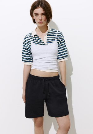 Ung person med kort brunt hår iført en stribet polo under en hvid camisoletop og sorte shorts med snor.