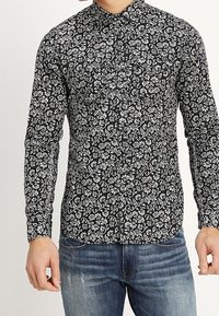 Uomo che indossa una maglietta nera attillata a maniche lunghe con motivo floreale bianco e jeans blu sbiaditi, in piedi con le braccia rilassate.