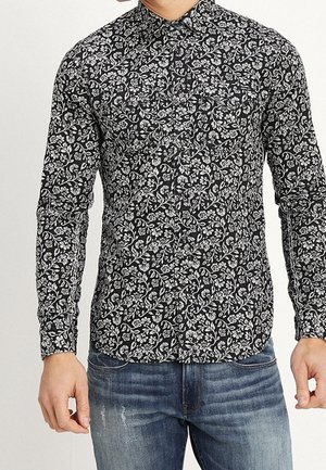 Homme portant une chemise noire ajustée à manches longues avec un motif floral blanc et un jean bleu délavé, se tenant avec les bras détendus.