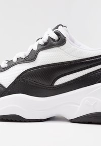 Musta-valkoinen urheilukenkä, jossa on teksturoitu päällinen, sileät yksityiskohdat ja paksu valkoinen pohja, jossa musta kuviointi. Nauhat ovat valkoiset.