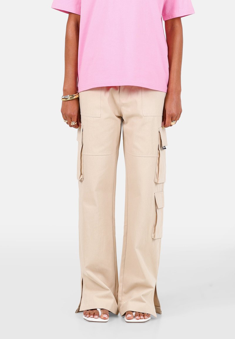 Person iført beige cargobukser med vide ben og sidelommer, lyserød kortærmet skjorte, hvide sandaler, ringe og armbånd, stående mod en hvid baggrund.