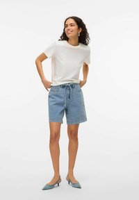 Lengvas baltas medvilninis marškinėliai, suderinti su mėlynais džinsiniais šortais, turinčiais reguliuojamą raištelį liemenyje. Modelis vilki šviesiai mėlynus aukštakulnius batus.