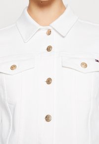 Giacca in denim bianco con bottoni brandizzati oro, tasche a pattina sul petto e un piccolo logo Tommy Hilfiger sulla tasca destra.