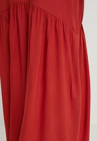 Czerwona sukienka z rozkloszowanym dołem, z marszczoną teksturą w okolicy biustu. Wykonana z miękkiej tkaniny o gładkim wykończeniu i zwiewnym kroju.