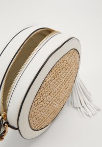 Kulatá taška s přední částí z tkaného slaměného materiálu, bílými texturovanými koženými stranami, zlatými zipy a dekorativní bílou třásní. Vhodná pro každodenní nošení.