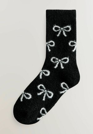 Sorte sokker med en fuzzy tekstur, der har et gentaget hvidt sløjfemønster overalt. Rund tådesign, velegnet til afslappet brug.