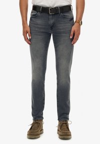 Vaqueros oscuros de hombre en azul marino con corte slim, con áreas desgastadas, cinco bolsillos y un cinturón negro. Combinados con zapatos de ante marrones.