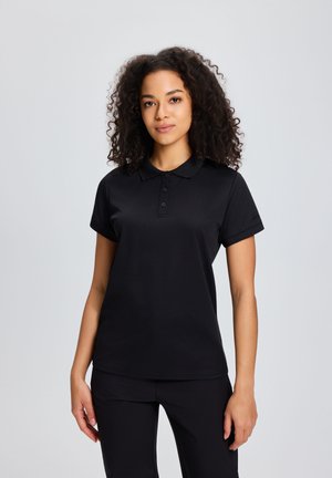 Młoda kobieta o kręconych włosach, ubrana w czarną koszulkę polo z krótkim rękawem i czarne spodnie, stojąca na tle jasnoszarego tła.