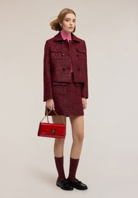 Burgundský tvídový sáčko a sukně s předními kapsami, sjednoceno s růžovým rolákem. Drží lesklou červenou kabelku se zlatými detaily.