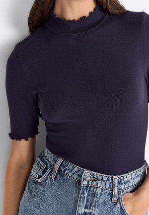 Frau trägt ein eng anliegendes, marineblaues, geripptes Kurzarmshirt mit Rüschenkragen und -ärmeln, in hoch taillierte, acid-wash blaue Jeans gesteckt.