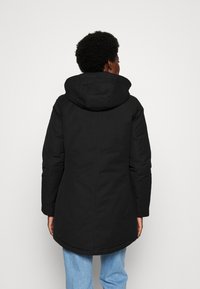 Cappotto isolato nero con cappuccio, caratterizzato da una linea dritta, texture liscia e orlo arrotondato. Indossato sopra dei pantaloni azzurri chiari.