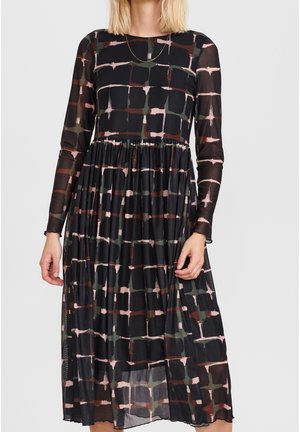 Długa czarna sukienka z siateczki z abstrakcyjnym wzorem w brązowo-oliwkową kratę, z marszczoną talią i długością do midi, noszona przez stojącą osobę.