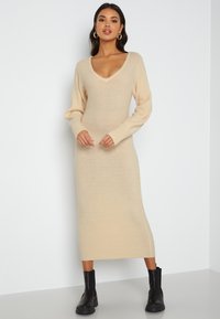 Abito a coste in beige con scollatura a V e maniche lunghe, caratterizzato da una silhouette aderente che arriva a metà polpaccio. Abbinato a stivaletti neri alla caviglia.