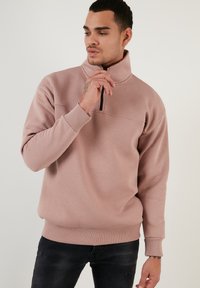 Sudadera de color rosa claro con un alto collar, media cremallera, puños y dobladillo acanalados, que presenta una textura suave y un corte casual.
